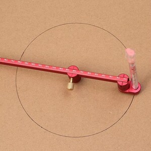 May include: A red compass tool with a ruler and a clear plastic tube is used to draw a circle on a brown surface. The compass has measurements in millimeters and a brass-colored adjustment knob.