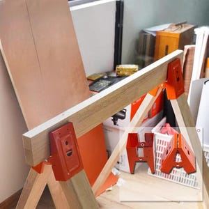 May include: A wooden sawhorse with orange metal brackets. The sawhorse has a light brown wooden beam and legs. The orange brackets connect the legs to the beam. The image also shows the letters "HA" in orange.