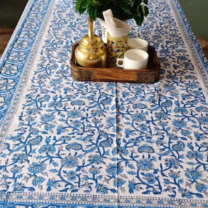 May include: A white and blue floral patterned tablecloth with a matching border. A wooden tray holds a brass teapot, two white cups, and a floral-decorated container with napkins. Green foliage adds a natural touch.