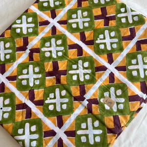 May include: A folded piece of fabric featuring a repeating geometric pattern of green squares with white cross motifs, set against a yellow background with dark purple intersecting lines and white diagonal stripes. A small tan tag displays the text 6.10.