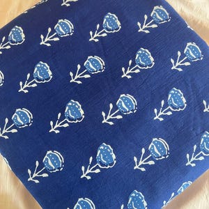 May include: A square piece of fabric in a deep blue color featuring a repeating pattern of white and light blue stylized flowers with stems and leaves. The textile has a visible woven texture and is folded into a neat, compact square shape.