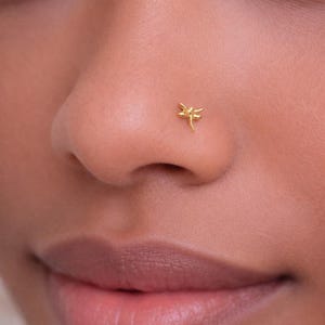 Op de afbeelding: Een close-up van een neus met een kleine, gouden libelle neusstud. De stud is gedetailleerd en toont de vleugels en het lichaam van de libelle. De huid heeft een warme tint en de lippen zijn zachtroze.