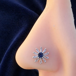 May include: A silver nose stud with a floral design. The centre of the flower is a dark blue gemstone, encircled by clear, baguette-cut stones. The jewellery is set against a light skin tone and a dark blue background.