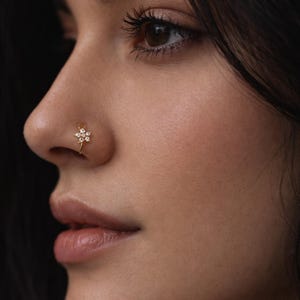 May include: A close-up of a person's face, with a gold nose ring. The nose ring has a floral design with small, clear gemstones. The skin tone is light, and the background is dark.