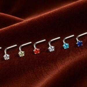 May include: A selection of nose studs, each featuring a star-shaped gemstone in shades of pink, clear, yellow, red, white, blue, dark blue, and purple. The studs are made of silver-coloured metal and are displayed on a brown velvet surface.