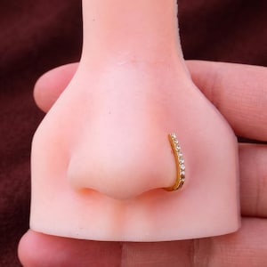 May include: A gold nose ring with a curved design, set with small, clear gemstones. The jewellery is displayed on a flesh-toned nose model, highlighting the ring's shape and sparkle.