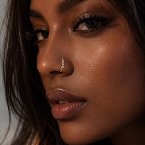May include: A gold nose ring featuring three small, clear, round-cut stones arranged in a vertical line. The jewellery is worn in the nostril, showcasing a thin, polished gold band that curves to fit the shape of the nose with a minimalist design.