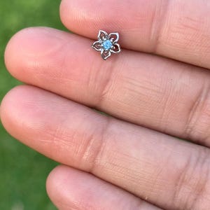 May include: A small, silver-coloured flower-shaped stud earring with a light blue centre stone. The earring is held between fingers against a blurred green background.