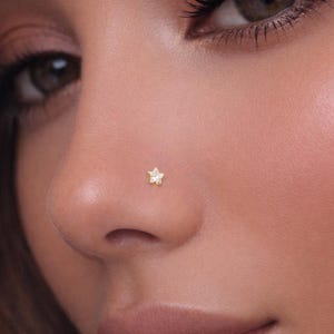 May include: Close-up of a nose with a small, gold-colored star-shaped nose stud. The stud is centered on the nostril. The skin tone is light with a soft, natural makeup look.