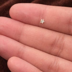 Puede incluir: Un pequeño pendiente en forma de estrella con un centro claro y brillante. El pendiente se sostiene entre los dedos, mostrando su delicado diseño. El fondo es una tela marrón suave, que proporciona un telón de fondo neutro para la joya.