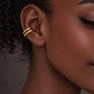 May include: Close-up of a gold ear cuff. The double-banded ear cuff is worn on the upper ear. The skin tone is dark, and the hair is curly. The jewelry is a simple, elegant design.