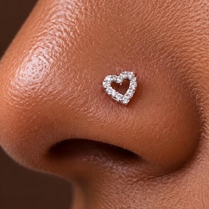 Puede incluir: Un piercing de nariz plateado con forma de corazón, engastado con pequeñas piedras transparentes y brillantes. La joya está situada en el lateral de la fosa nasal, mostrando un acabado metálico delicado y reflectante con múltiples cristales.