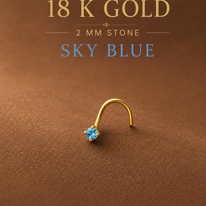 May include: An 18 K gold nose stud featuring a 2 mm sky blue stone. The text 18 K GOLD, 2 MM STONE, and SKY BLUE is displayed above the jewelry. The stud has a curved post and a four-prong setting holding the round, faceted blue gemstone.