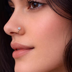 May include: A close-up of a person's face, featuring a silver nose ring with a small, sparkling, flower-shaped design. The nose ring is set against the person's skin and dark hair.