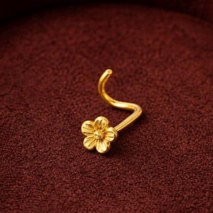 Op de afbeelding: Een goudkleurige neusring met een bloemmotief. De neusring heeft een gebogen staaf en een kleine, gedetailleerde bloem aan het uiteinde. De bloem heeft vijf bloemblaadjes en een getextureerd midden.