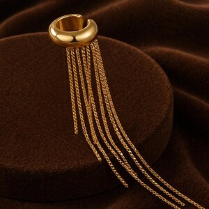 May include: A gold-colored ear cuff with a rounded design and multiple dangling chain strands. The jewelry piece is set against a brown background, creating a contrast that highlights the ear cuff's metallic shine.