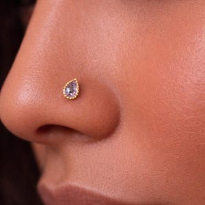 May include: Close-up of a gold nose stud with a teardrop-shaped, light purple gemstone. The stud is set in a gold bezel with a beaded edge, and is placed on a person's nose.