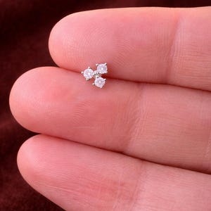 Puede incluir: Un pequeño pendiente de botón plateado con tres piedras de circonita cúbica redondas y transparentes dispuestas en un racimo triangular. El pendiente se muestra sobre un fondo borroso de color marrón rojizo oscuro.