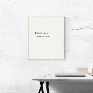 May include: A framed print with the text "This is not a conversation" in a minimalist style. The print has a cream-colored background and a thin gold frame. The print is on a white desk with a laptop and other items.