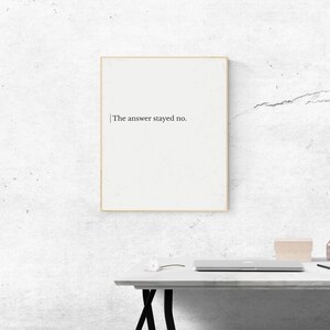 Op de afbeelding: Een ingelijste print met de tekst "The answer stayed no." Het kunstwerk heeft een minimalistisch ontwerp met zwarte tekst op een witte achtergrond, omlijst door een dunne gouden lijst. De print wordt weergegeven op een witte tafel tegen een getextureerde muur.