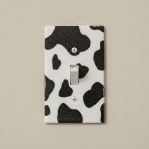 May include: A decorative light switch cover with a black and white cow print pattern. The switch plate has a white background with black irregular spots. The light switch is a light gray color.