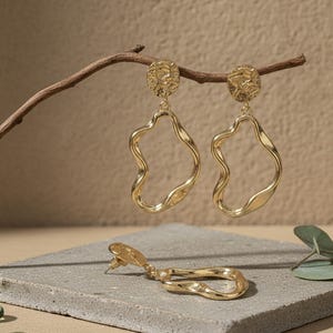 Puede incluir: Pendientes dorados con una parte superior circular texturizada y una gota ondulada irregular. Un pendiente descansa sobre una superficie gris, mientras que dos cuelgan de una rama marrón. Los pendientes tienen un acabado pulido y un diseño moderno y abstracto.