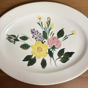 May include: Oval, off-white ceramic platter with a hand-painted floral design. The flowers include yellow, pink, and purple blooms with green leaves. The rim has a yellow dotted border. A decorative serving piece.