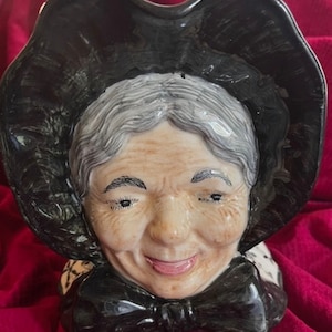 May include: A ceramic figurine of a woman's head wearing a black bonnet and a black bow. The woman has gray hair and a smiling face.