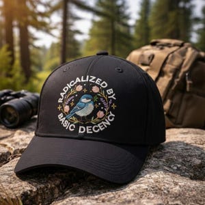 Op de afbeelding: Zwarte baseballcap met de geborduurde tekst "RADICALIZED BY BASIC DECENCY" rond een blauwe vogel. De cap ligt op een rots met een vage bosachtergrond.