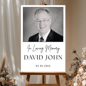 Puede incluir: Un letrero conmemorativo con una foto en blanco y negro de un hombre con traje y gafas. El letrero dice "In Loving Memory", el nombre "DAVID JOHN" y una fecha.