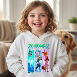 Peut inclure: Un sweat &agrave; capuche blanc avec le texte "ZOOTOPIA 2" en vert et bleu. Le sweat &agrave; capuche pr&eacute;sente un graphisme des personnages du film Zootopia. Une jeune fille porte le sweat &agrave; capuche.