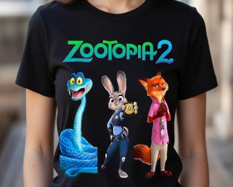 Peut inclure: T-shirt noir avec des personnages de Zootopie 2. Le motif comprend un serpent bleu, un lapin en uniforme de policier et un renard en chemise rose. Les mots "ZOOTOPIA 2" sont imprim&eacute;s en vert et bleu en haut.
