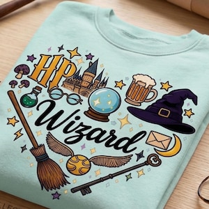 May include: A light mint green sweatshirt featuring a graphic print with the text "Hp Wizard" surrounded by illustrations of a castle, a crystal ball, a witch hat, a broom, a golden snitch, a key, a mug, glasses, mushrooms, and an envelope.