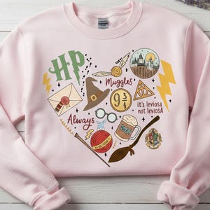 May include: A light pink sweatshirt featuring a heart-shaped design with various illustrations and text. The design includes a lightning bolt, a Hogwarts crest, a golden snitch, a sorting hat, and the words "Always" and "Muggles".