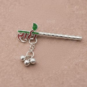 May include: A silver-colored flute-shaped pendant with the word "राधा" in red and green lettering. The pendant has a chain with three small silver bells attached. The background is a solid, light brown color.
