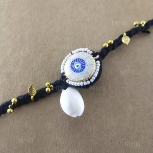 May include: A black braided bracelet featuring a central circular charm with a blue and white eye design. The bracelet is adorned with gold-colored beads and leaf-shaped charms, and a white seashell pendant.