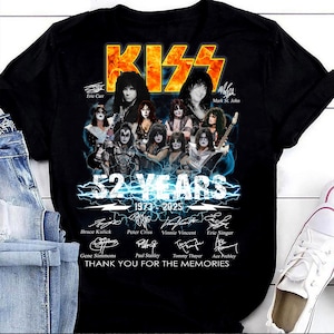 Peut inclure: T-shirt noir avec le groupe KISS et le texte "KISS" en orange et jaune. L'image présente les membres du groupe, le texte "52 YEARS 1973-2025", des signatures et la phrase "THANK YOU FOR THE MEMORIES".