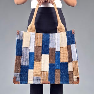 May include: A large tote bag with a patchwork design in shades of blue, brown, and beige. The bag has woven handles and leather corner accents. The bag is held by a person.