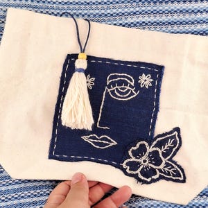 以下が含まれることがあります： クリーム色のトートバッグ。ネイビーブルーの正方形に顔の刺繍、花、タッセルが施されています。バッグにはネイビーブルーの紐と黄色のビーズが付いています。