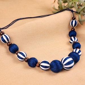 May include: A handmade necklace with alternating navy blue and white striped spheres and solid navy blue spheres. The necklace is strung on a dark blue cord, with small wooden beads between the spheres. The necklace is displayed on a beige surface.