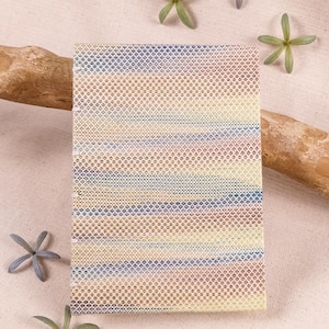 Op de afbeelding: Een rechthoekige, geweven placemat met een gradiëntpatroon van geel, bruin, blauw en wit. De placemat is gemaakt van een textuurrijk materiaal en is omgeven door kleine groene en grijze stervormige bloemen en een stuk drijfhout.