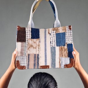 Può includere: Una borsa tote patchwork con una varietà di motivi in tessuto nei toni del blu, marrone e crema. La borsa ha una forma rettangolare con angoli in pelle marrone e manici robusti. La borsa è tenuta da una persona.