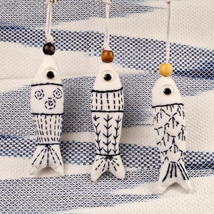 May include: Three white fish-shaped keyrings with black embroidered details and wooden beads. Each keyring is suspended from a silver ring and hangs against a blue and white patterned background. The fish have different embroidered patterns.
