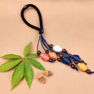 Peut inclure: Un élastique à cheveux noir avec une boucle, orné d'un groupe de perles et de pompons colorés dans les tons rose, orange, blanc, bleu et jaune. L'image comprend également des feuilles vertes et de petites gousses.