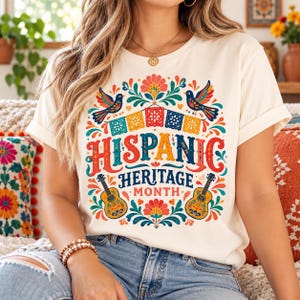 May include: A cream-colored t-shirt featuring a colorful graphic print with the text "HISPANIC HERITAGE MONTH". The design includes two birds, floral patterns, decorative banners, and two acoustic guitars in shades of red, blue, yellow, and green.