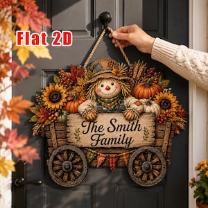 May include: A decorative door sign featuring a scarecrow sitting in a wooden wagon with wheels, surrounded by sunflowers, pumpkins, and autumn leaves. The sign displays the text 'The Smith Family' in dark script. It hangs from a rope.