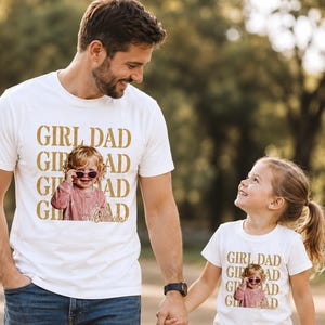 Pu&ograve; includere: Una maglietta bianca con la scritta "GIRL DAD" in lettere dorate, con una foto di una bambina con gli occhiali da sole. L'immagine mostra un padre e sua figlia che si tengono per mano, entrambi con magliette abbinate.