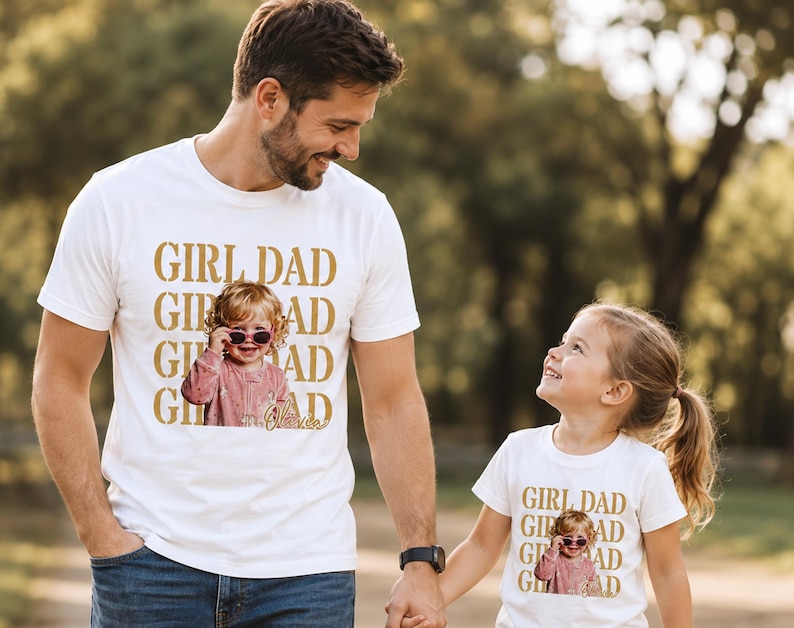 Pu&ograve; includere: Una maglietta bianca con la scritta "GIRL DAD" in lettere dorate, con una foto di una bambina con gli occhiali da sole. L'immagine mostra un padre e sua figlia che si tengono per mano, entrambi con magliette abbinate.