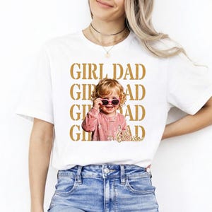 Pu&ograve; includere: T-shirt bianca con le parole "GIRL DAD" in lettere dorate. Una foto di un bambino sorridente con occhiali da sole &egrave; centrata sulla maglietta. Il bambino indossa un completo rosa. Il nome "Olivia" &egrave; scritto in corsivo.