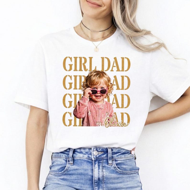 Pu&ograve; includere: T-shirt bianca con le parole "GIRL DAD" in lettere dorate. Una foto di un bambino sorridente con occhiali da sole &egrave; centrata sulla maglietta. Il bambino indossa un completo rosa. Il nome "Olivia" &egrave; scritto in corsivo.